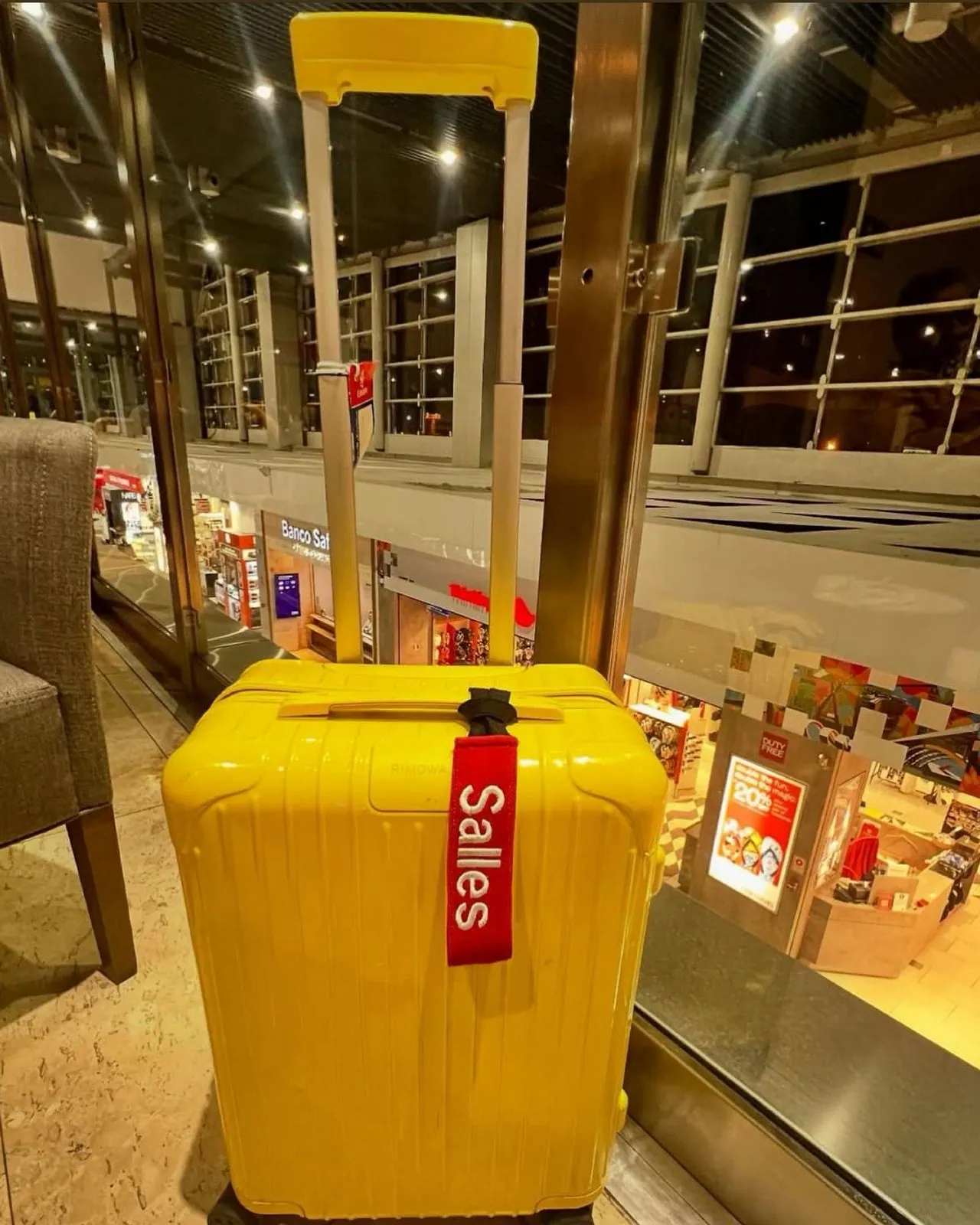 Tag vermelha de elástico presa na alça de uma mala amarela, na área de free shop de um aeroporto.