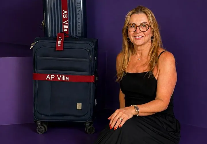 Paula, fundadora da marca, sentada e sorrindo ao lado de duas malas azul-marinho com faixas e tag vermelhas bordadas.